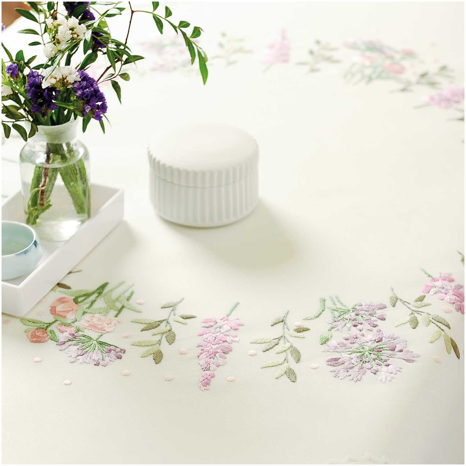 Stickpackung Tischdecke Blumenkranz 90x90cm zeigt zarte Blumenstickereien auf cremefarbenem Stoff, dekoriert mit einem Blumenstrauß in einer Vase.