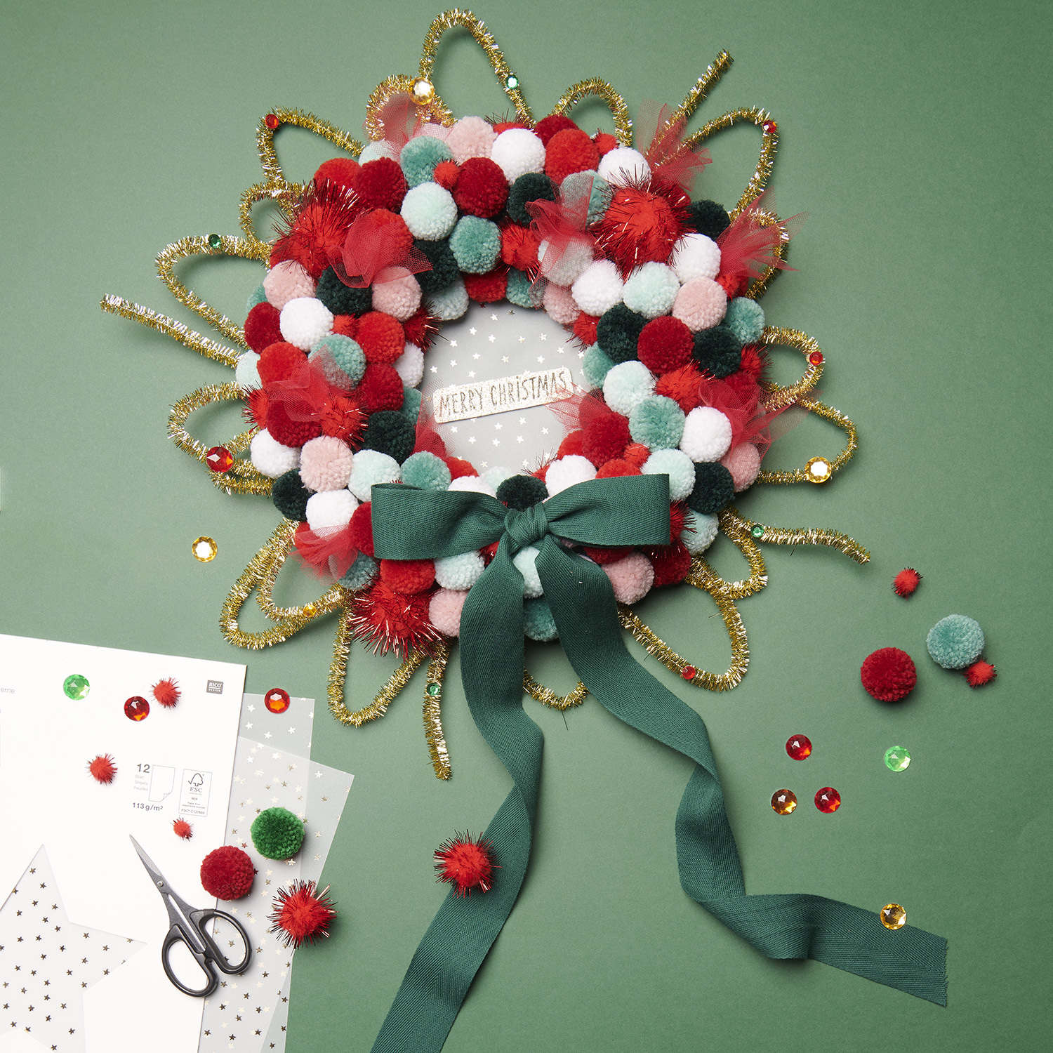 Bastelanleitung Weihnachtskranz mit Pompons: Bunter Kranz aus roten, grünen und weißen Pompons, verziert mit Schleife und Glitzer auf grünem Hintergrund.