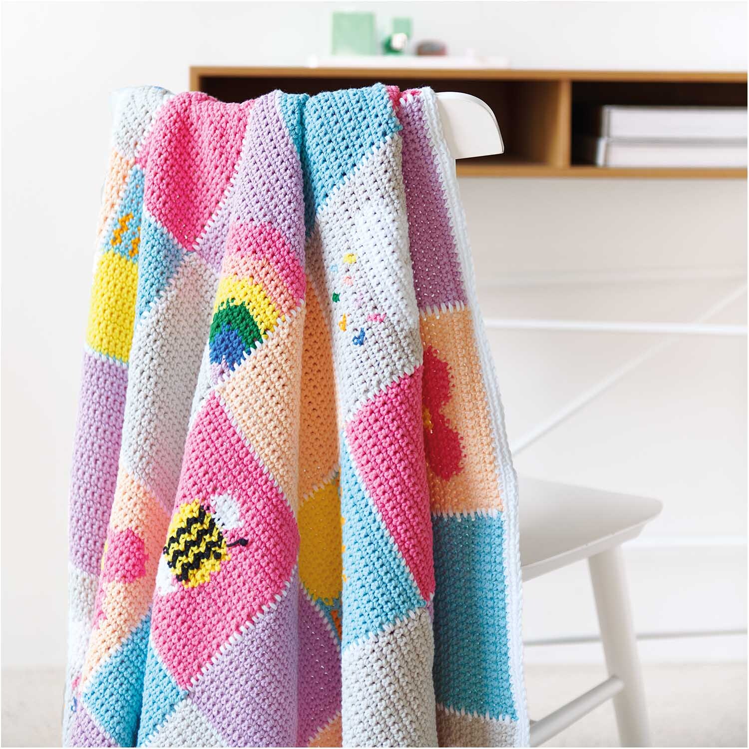 Creative Cotton aran: Bunte, gehäkelte Decke mit geometrischen Mustern auf einem weißen Stuhl.