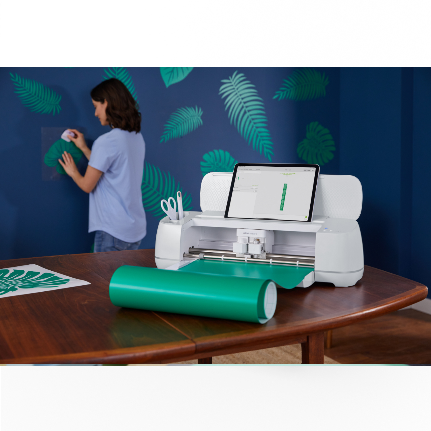 Maker 3 Schneideplotter von Cricut auf einem Tisch, während eine Person grüne Blätter an die Wand klebt.