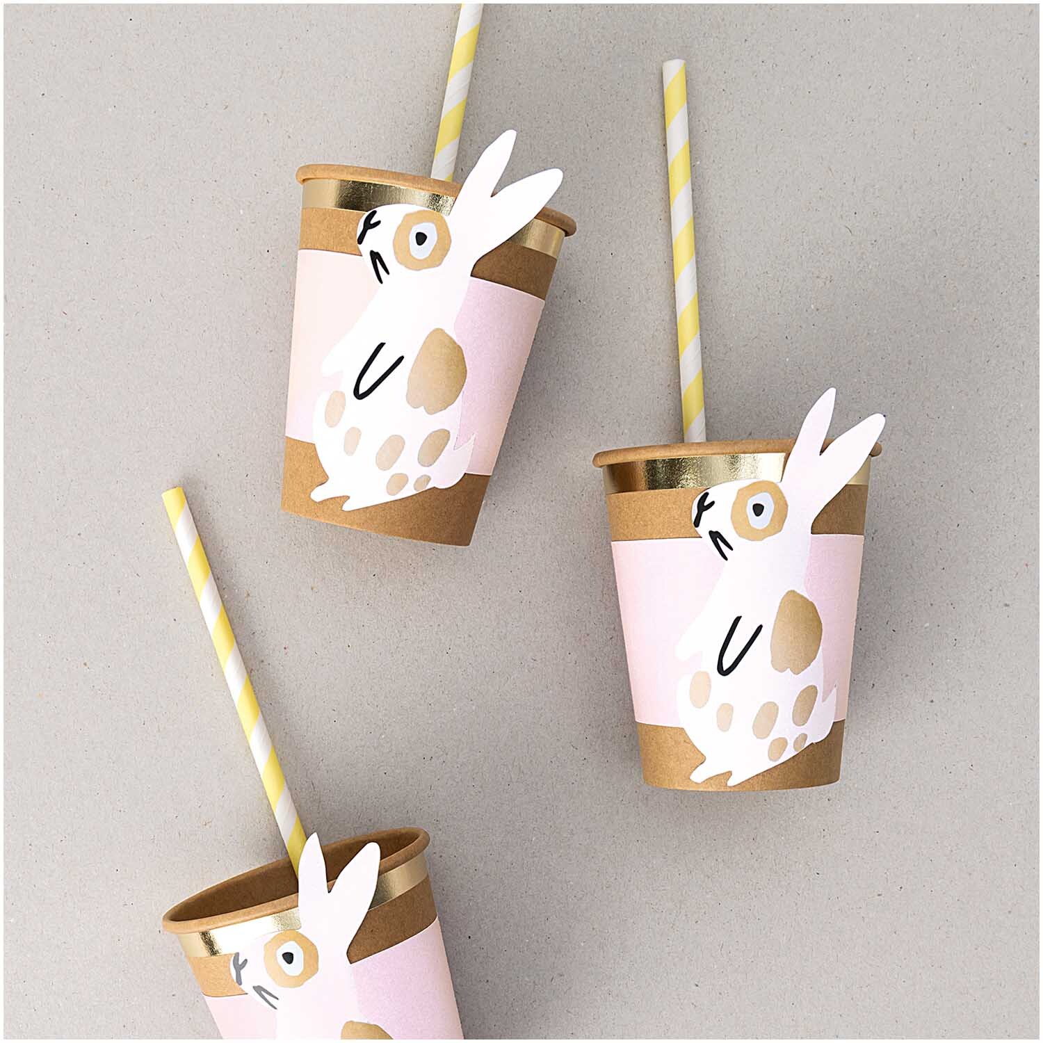 Papiermanschetten Hase rosa 6 Stück: Drei rosa Becher mit Hasenmotiv und gelben Strohhalmen auf grauem Hintergrund.