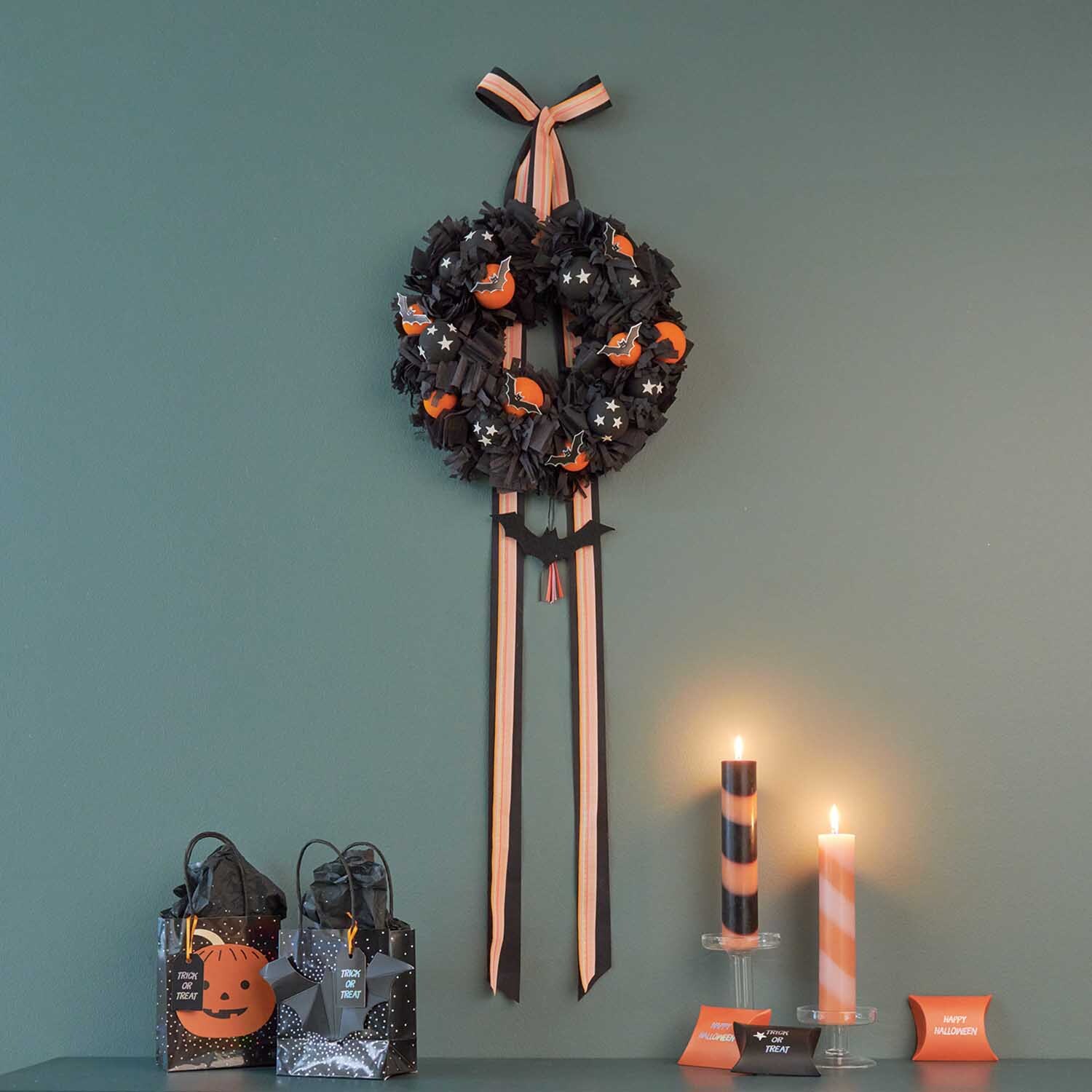 Bastelanleitung Halloween-Türkranz: Schwarzer Kranz mit orangefarbenen Akzenten, dekoriert mit Schleifen, an grüner Wand.