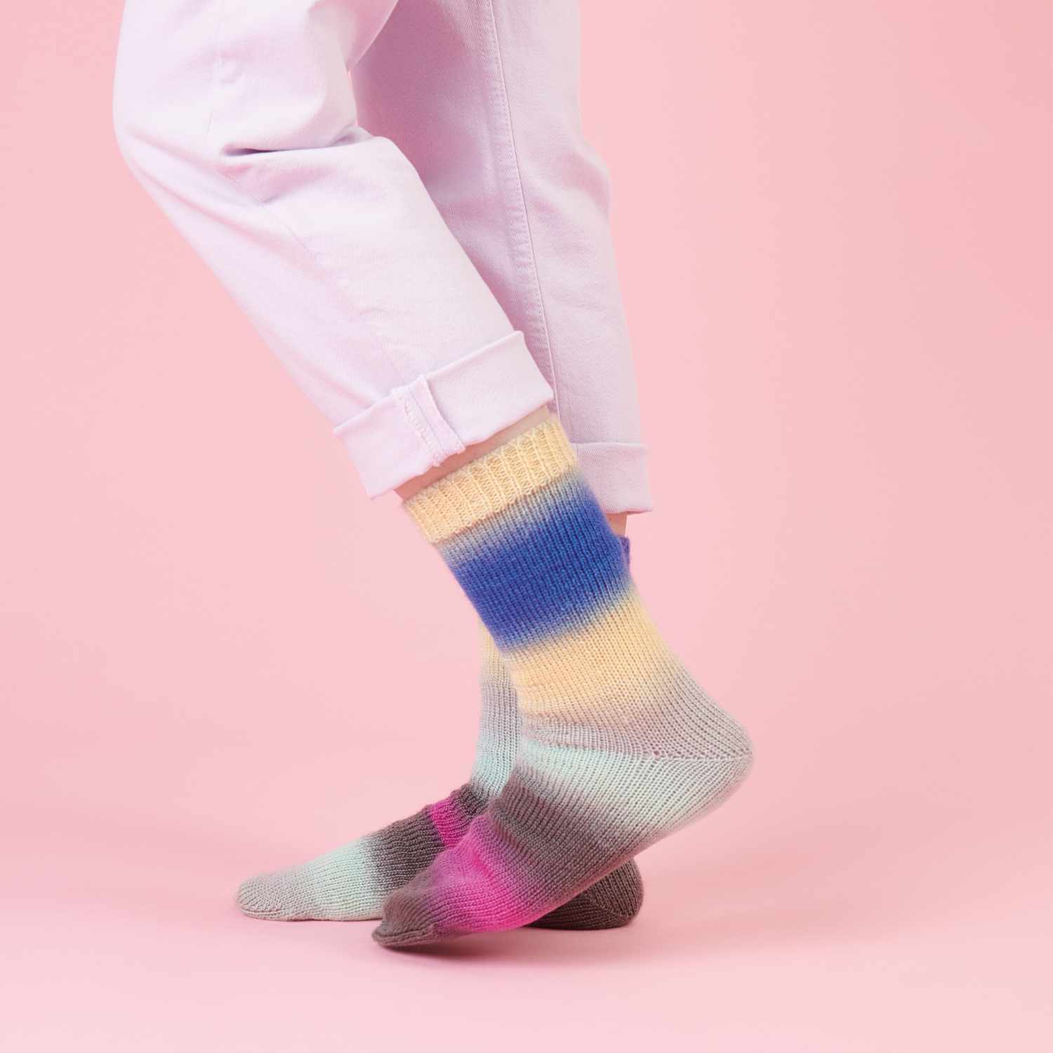 Superba Twirl 4fädig: Bunte gestrickte Socken in Pastelltönen auf rosa Hintergrund, getragen von Person in hellen Hosen.