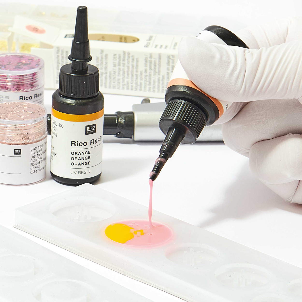 Hand, welche rosa UV-Resin in eine Silikonform gießt, dahinter Blattmetall und ein UV-Resin-Fläschchen.