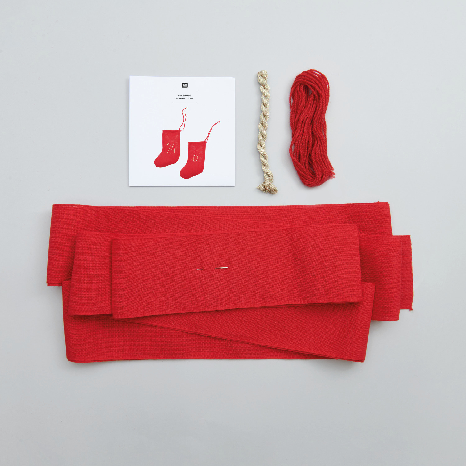 Stickpackung Stiefel-Adventskalender von Rico Design mit rotem Stoff, Garn und Anleitung auf grauem Hintergrund.