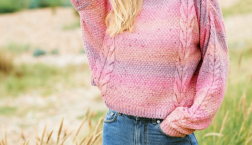 Strickanleitung Pullover aus Fashion Cotton Light & Long Tweed dk: Frau trägt rosa Pullover mit Zopfmuster, kombiniert mit blauen Jeans in natürlicher Umgebung.