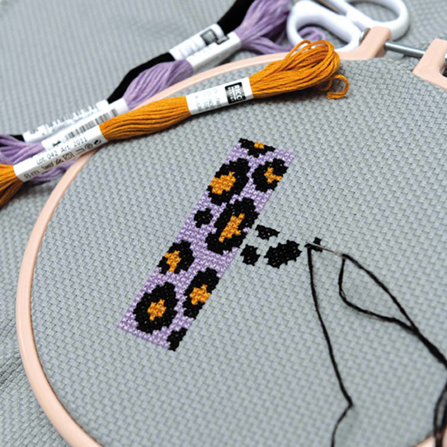 Sticktwist 8m von Rico Design: Ein Stickrahmen mit Leopardenmuster in Lila und Orange auf grauem Stoff.