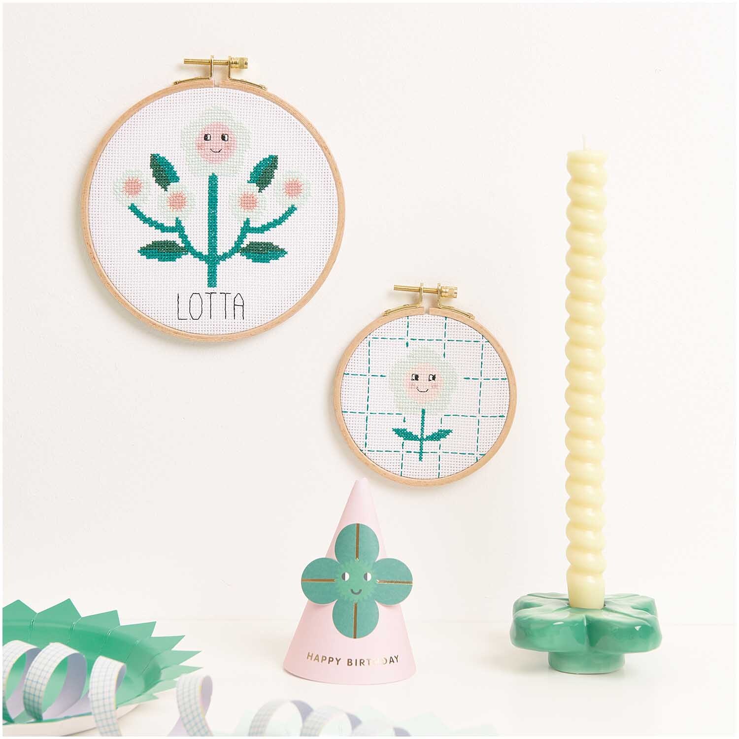 Stickpackung Happy Flower von Rico Design zeigt zwei bestickte Blumenrahmen mit Kerze und Dekoration.