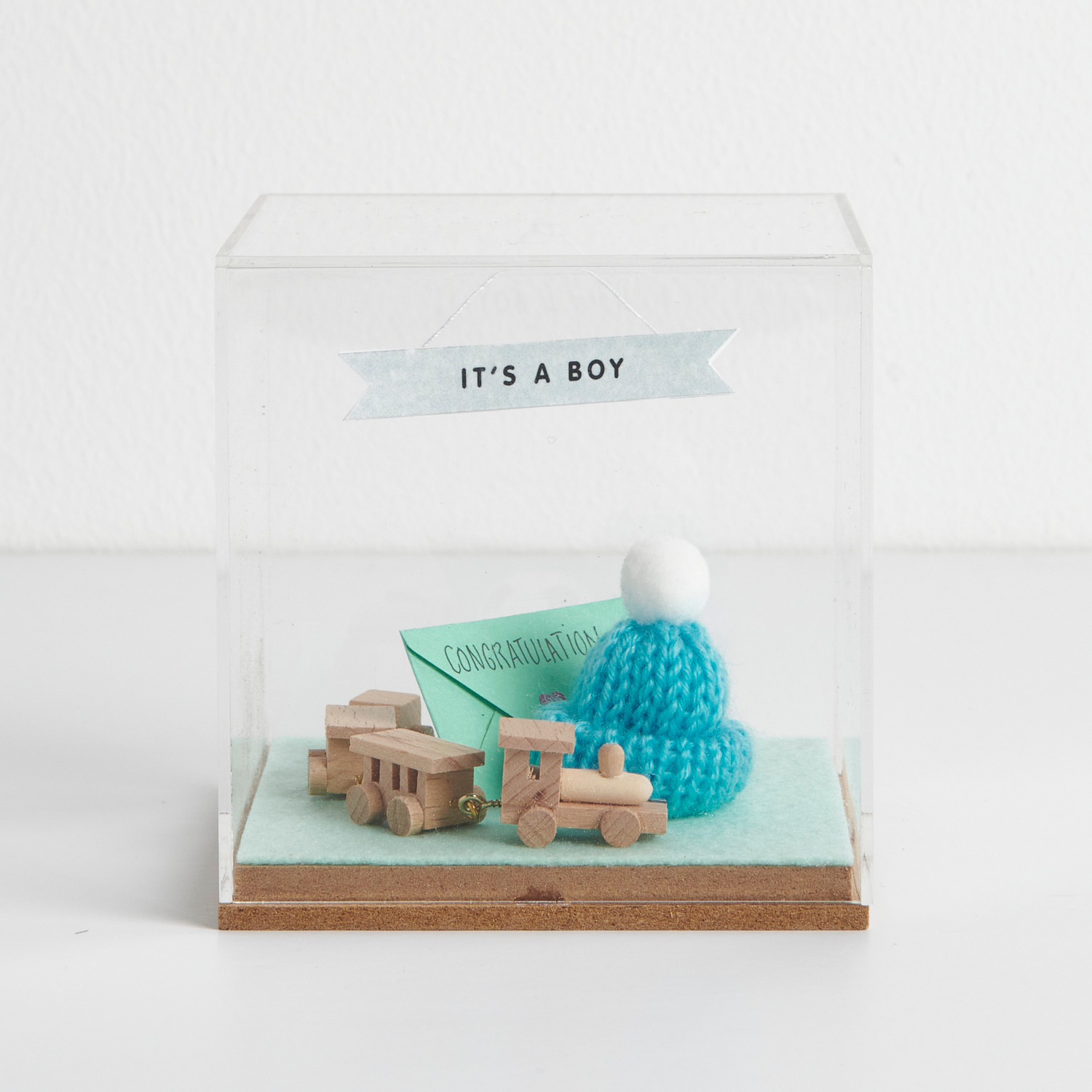 Figurico Dekohaube Würfel mit Holzspielzeugzug, blauem Strickmütze und "It's a Boy"-Banner. Ideal als Geschenk zur Geburt.