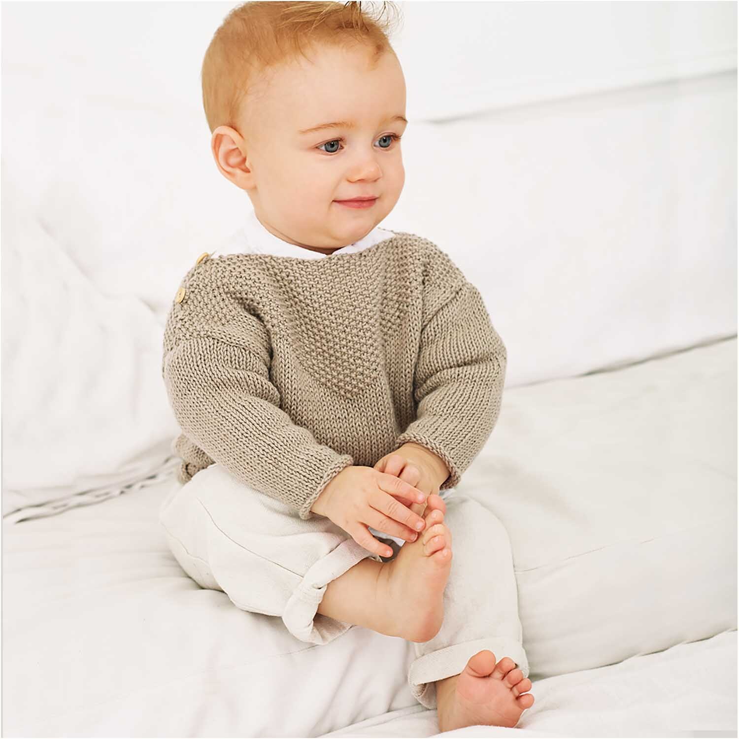 Rico Baby Cotton Soft dk: Ein Baby trägt einen gestrickten Pullover in Oliv, sitzt auf einem weißen Bett.