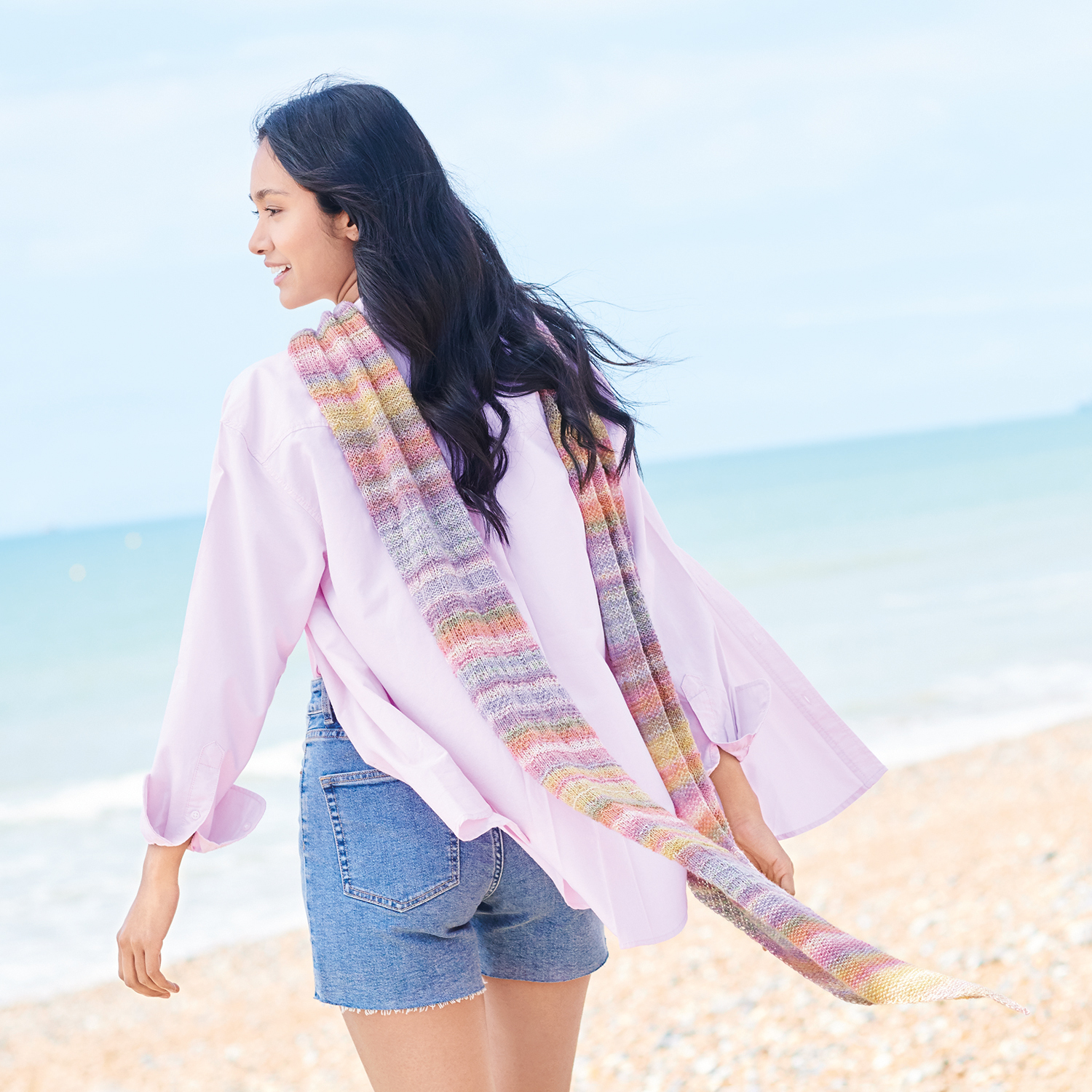 Strickanleitung Tuch aus Fashion Cotton Merino Lace: Frau am Strand mit buntem Schal, lila Hemd und Jeansshorts.