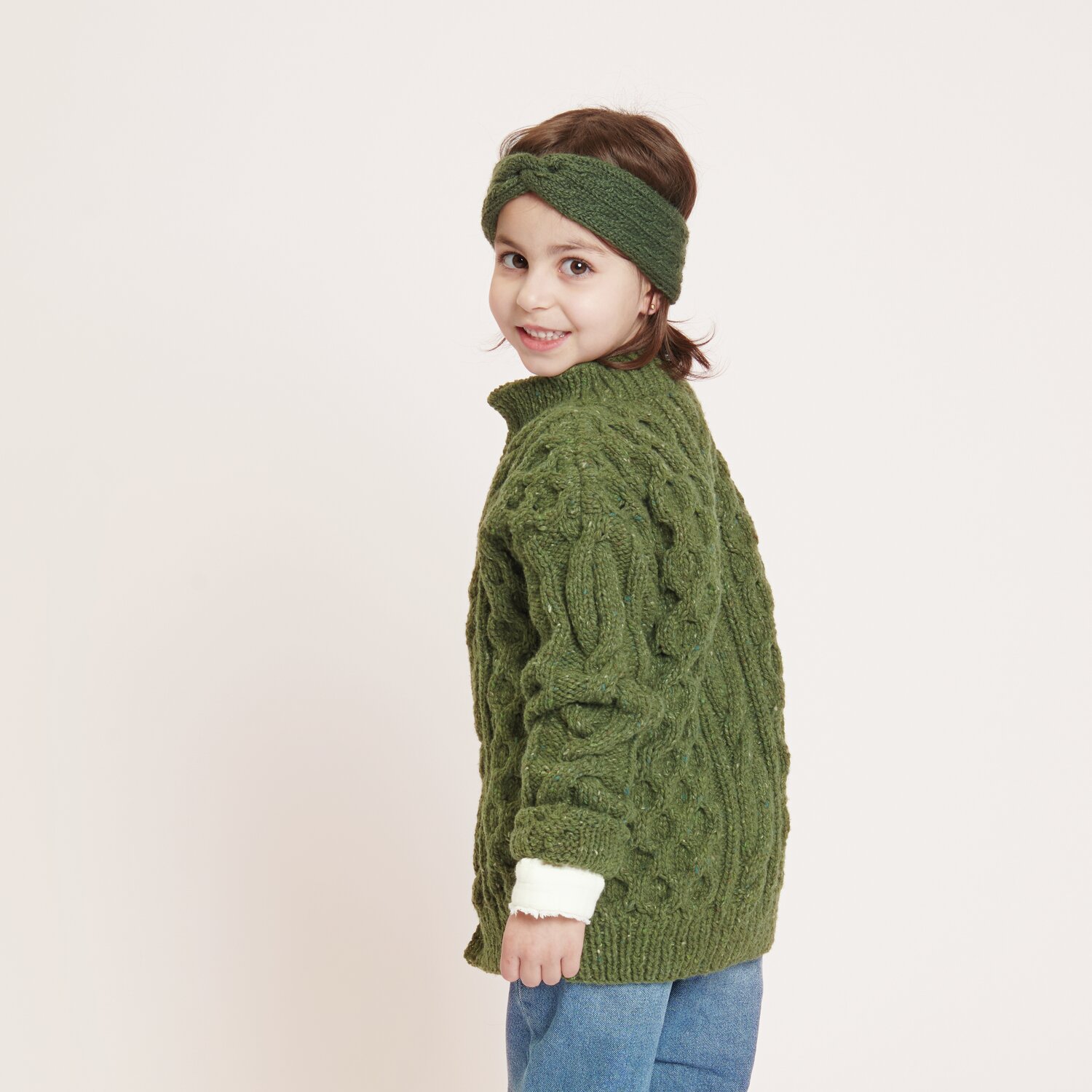 Strickset Kinderjacke Modell 24: Kind in grüner Strickjacke und Stirnband, lächelnd zur Seite blickend.