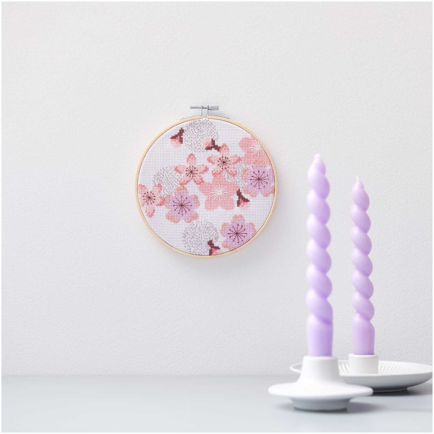 Stickpackung Kirschblüten Bild Ø 15cm zeigt zarte rosa Blüten in einem Stickrahmen an einer weißen Wand.