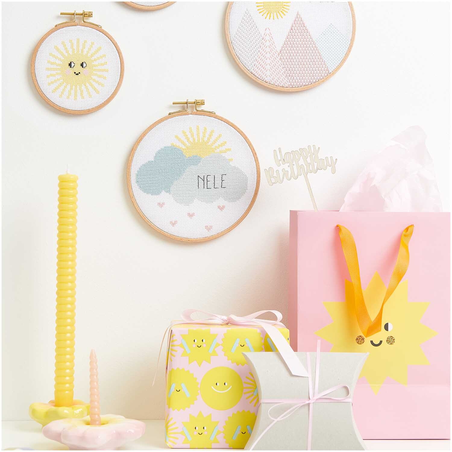 Stickpackung Geburtsbild Wolken von Rico Design zeigt bunte Stickrahmen mit Wolkenmotiv, umgeben von fröhlichen Geschenken und Dekorationen.