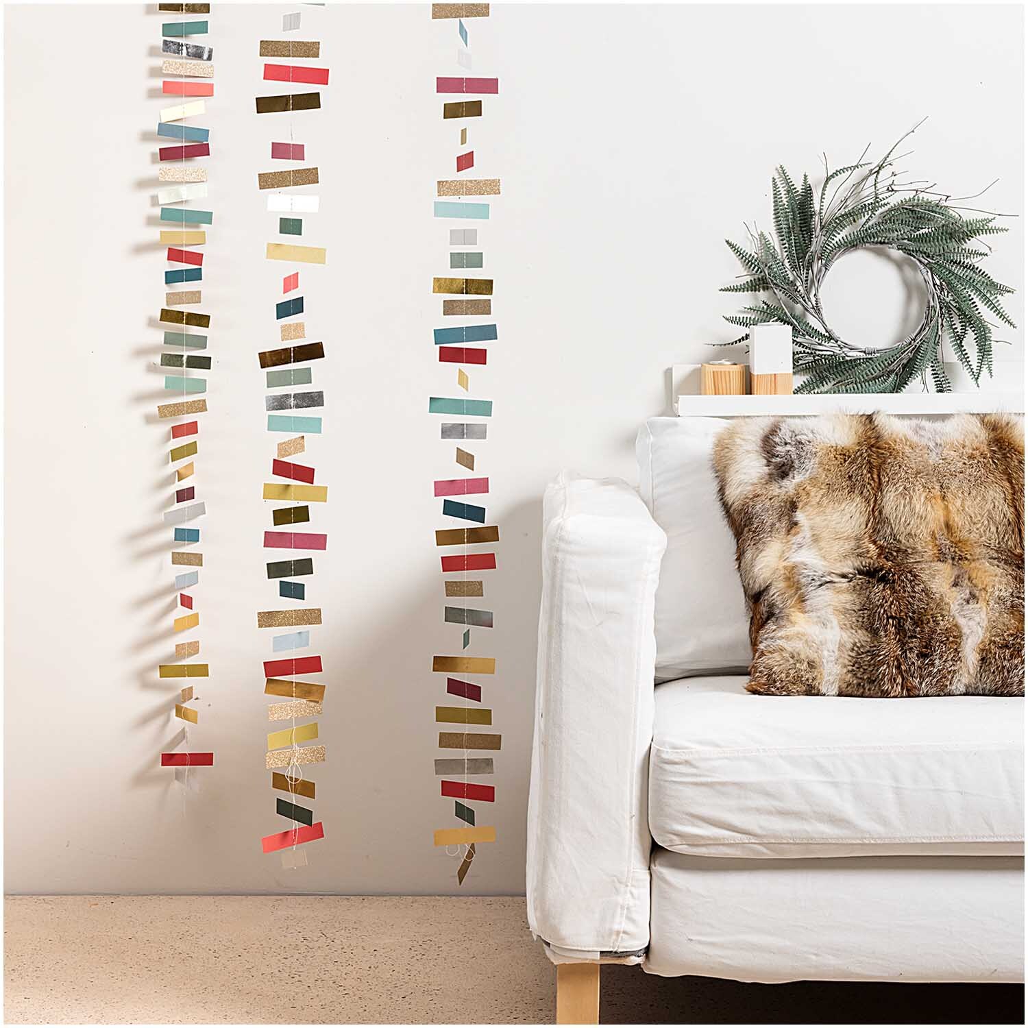 Genähte Girlande Streifen classic 1,8m von Rico Design in bunten Farben, dekorativ an einer weißen Wand neben einem Sofa.