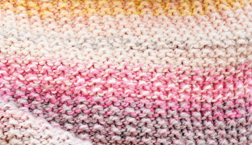 Strickanleitung Dreieckstuch aus Chic-Unique: Nahaufnahme eines gestrickten Tuchs in sanften Rosa- und Gelbtönen.