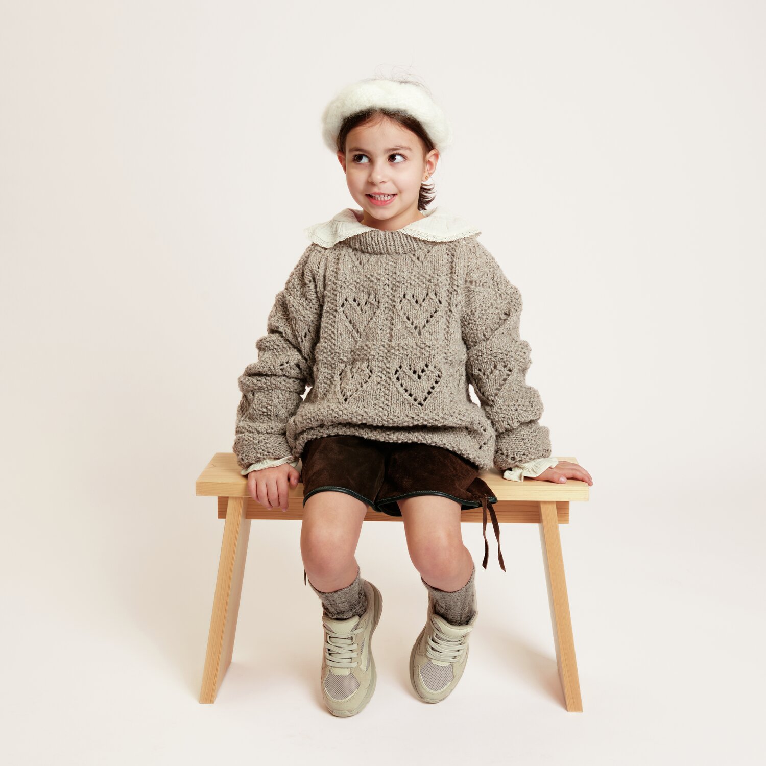 Strickset Kinderpullover Modell 06: Kind in grauem Pullover mit Herzmuster, sitzt auf Holzbank.