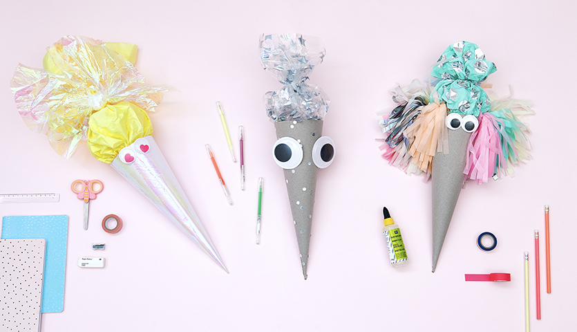 Bastelanleitung Geschwister-Schultüten mit Wackelaugen: Drei bunte Schultüten mit Augen, umgeben von Bastelmaterialien auf rosa Hintergrund.