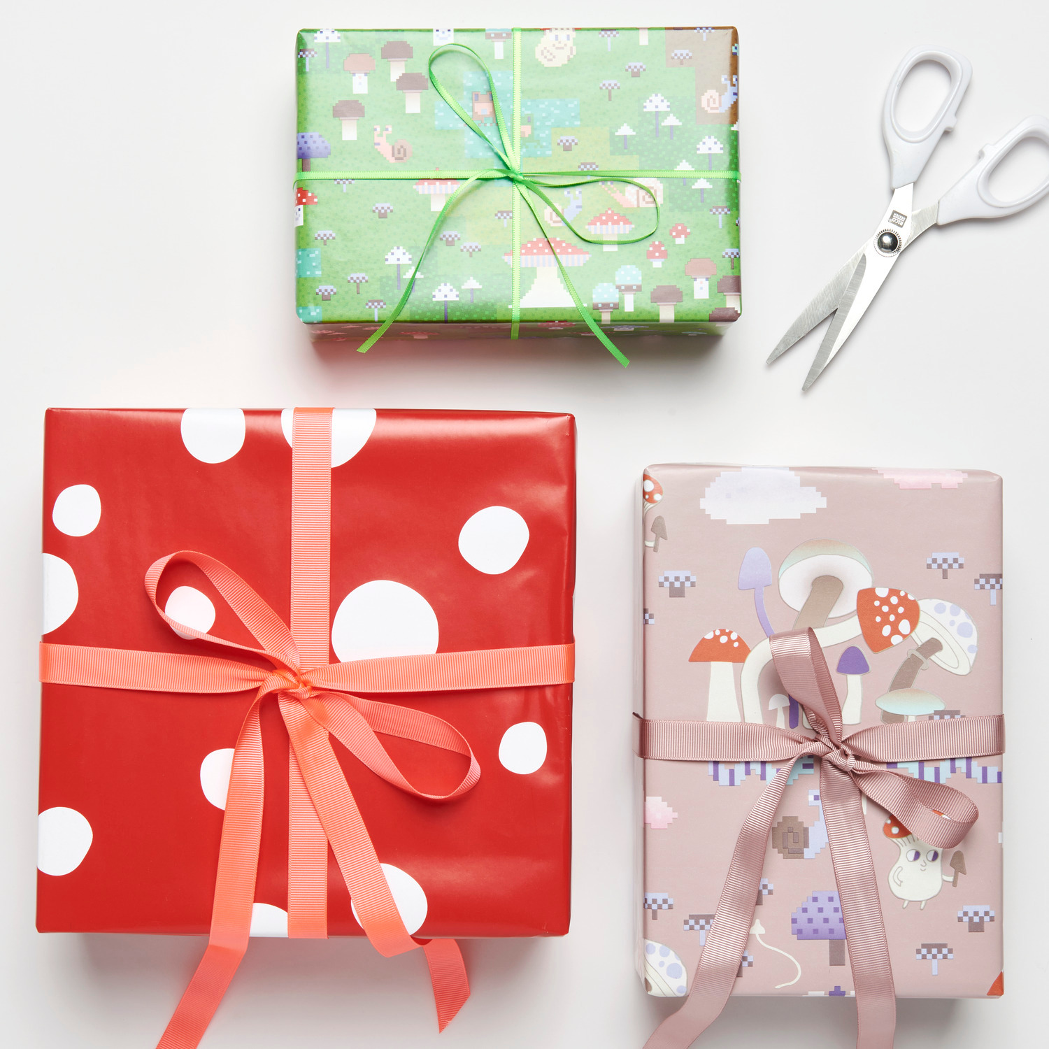 Geschenkpapier Shrooom Games, Fliegenpilz-Muster in Rot, Grün und Rosa, dekorativ verpackt mit Schleifen, daneben eine Schere.