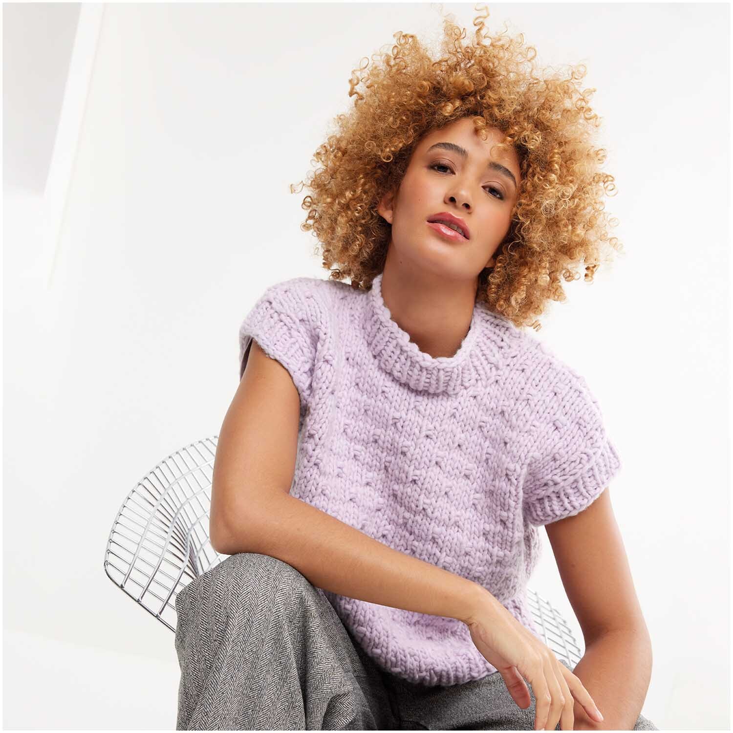 Strickset Pullunder Modell 02: Frau trägt lila Pullunder, sitzt auf Stuhl, moderne Mode.