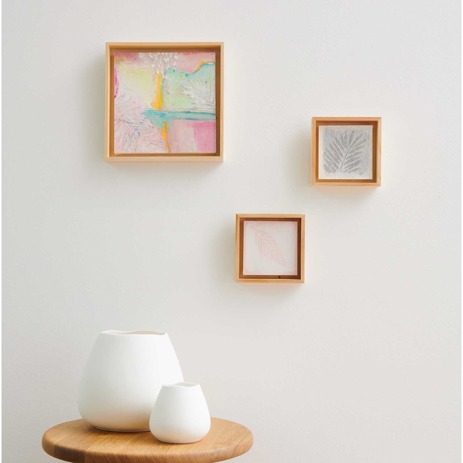 Ölpastellkreide 36 Farben von Rico Design: Drei gerahmte Kunstwerke an einer weißen Wand über zwei weißen Vasen auf einem Holztisch.