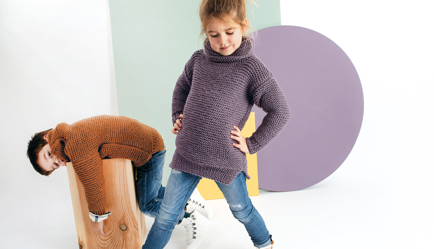 Strickanleitung Rollkragenpullover für Kinder: Zwei Kinder in gestrickten Pullovern, eines in Lila, das andere in Braun, posieren spielerisch vor geometrischen Formen.