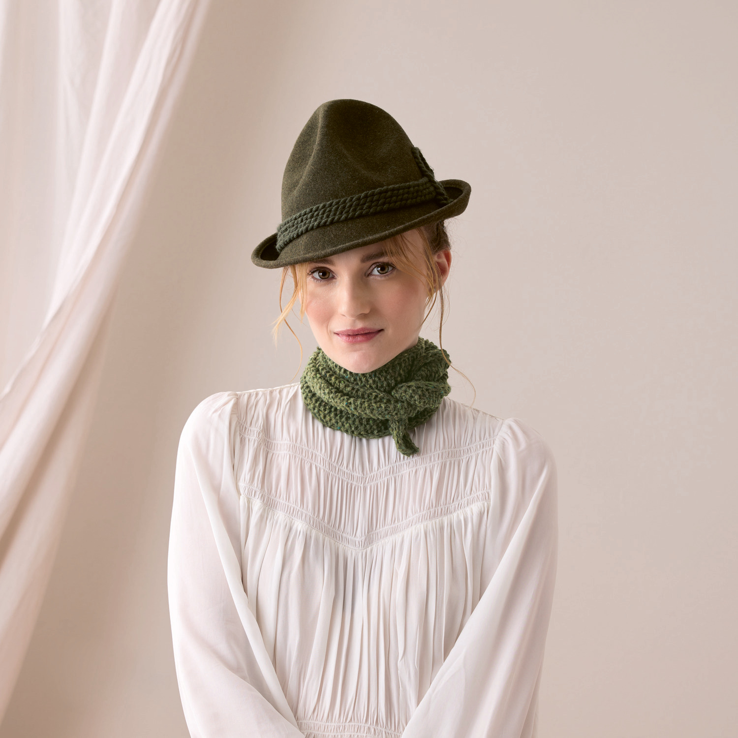 Strickset Tuch Modell 05: Frau in weißer Bluse mit olivgrünem Schal und Hut von Rico Design.