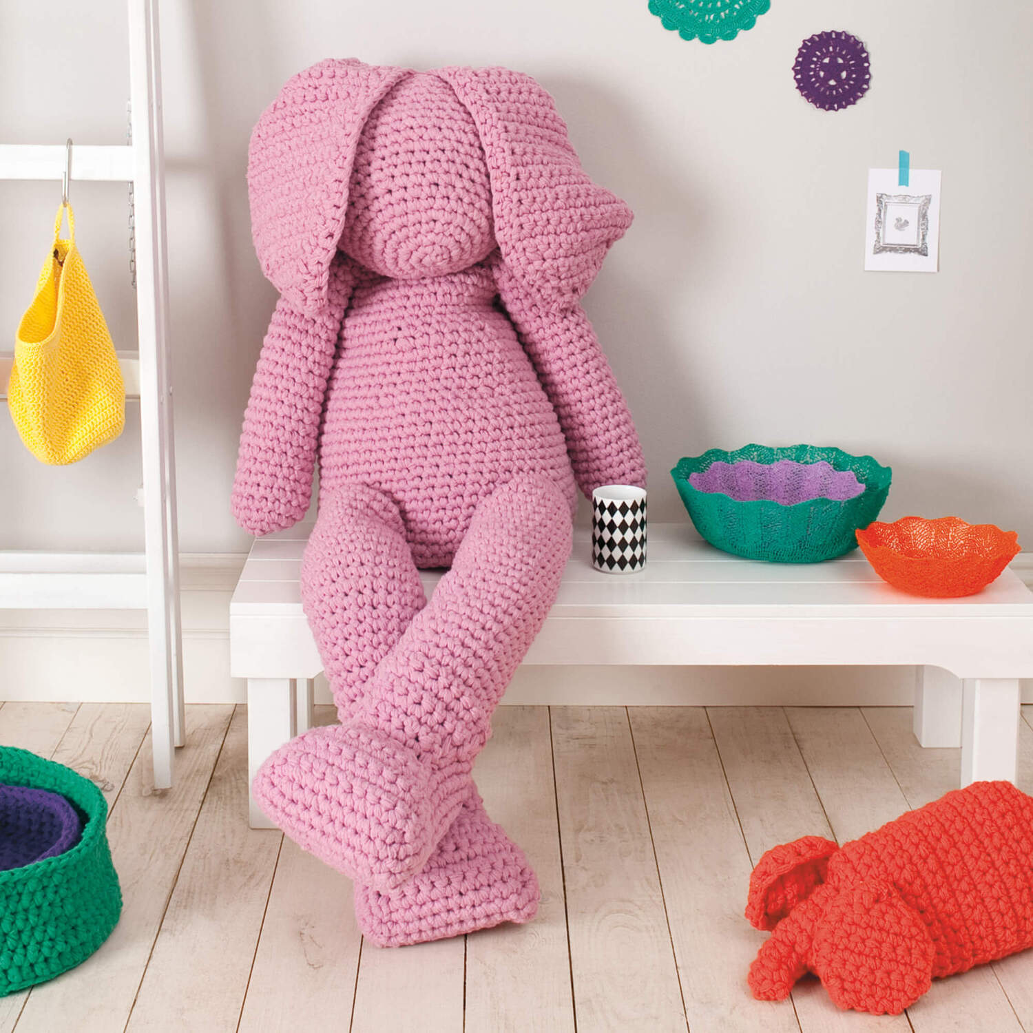 Häkelanleitung XXL Hase aus Creative Loopy: Ein großer, rosa gehäkelter Hase sitzt auf einer Bank, umgeben von bunten Körben.
