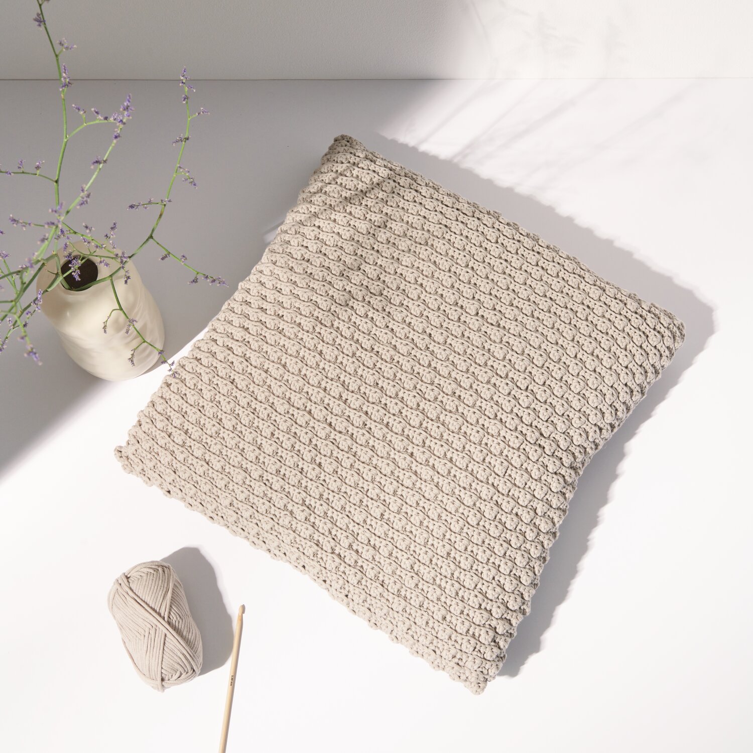 Häkelset Kissen Modell 07 aus Boho Crochet: Beiges Kissen mit Häkelmuster, Garnknäuel und Häkelnadel auf weißem Hintergrund.