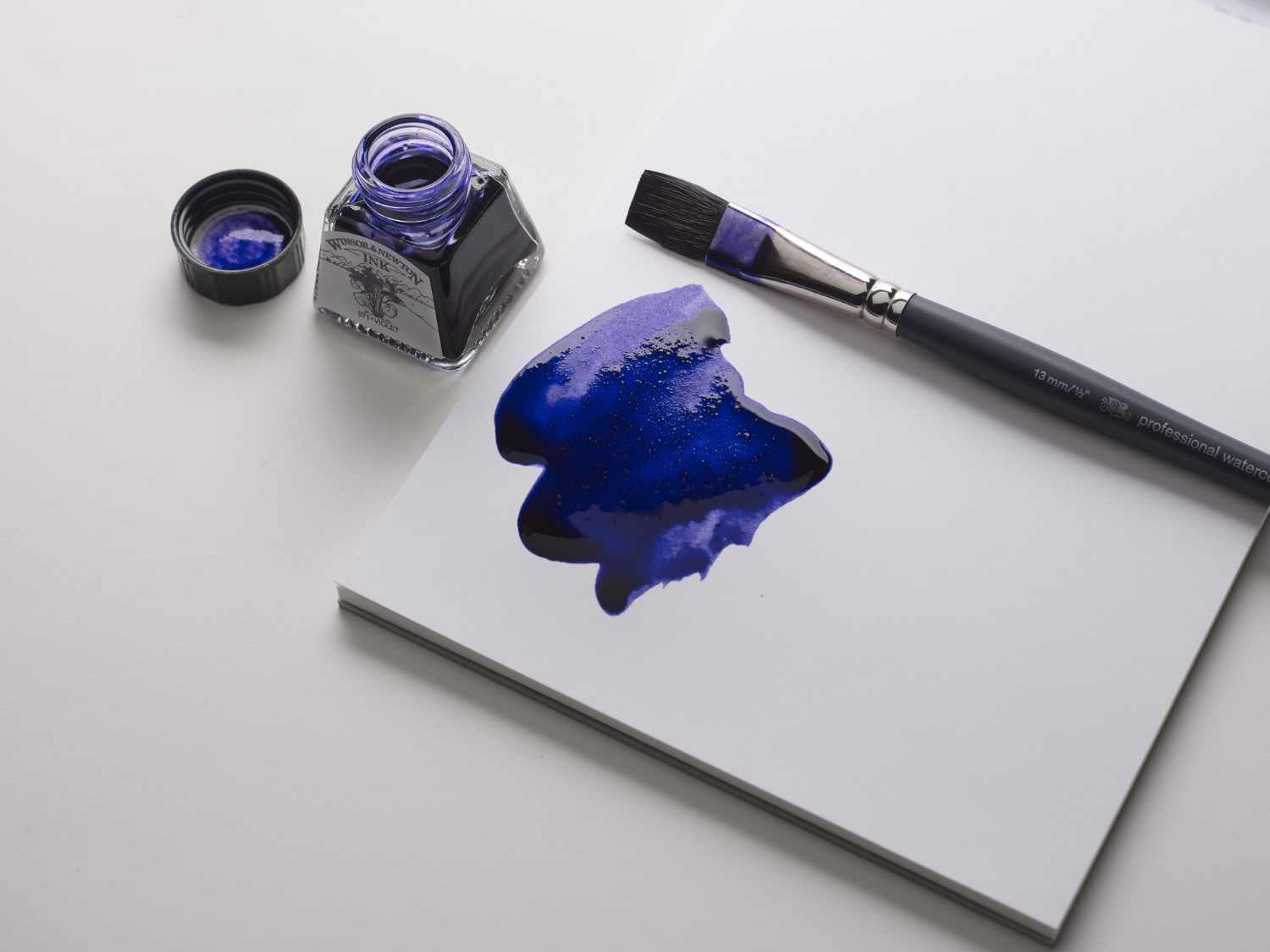 Zeichentusche 14ml von Winsor & Newton in Violett, offen mit Pinsel und Tuschfleck auf weißem Papier.