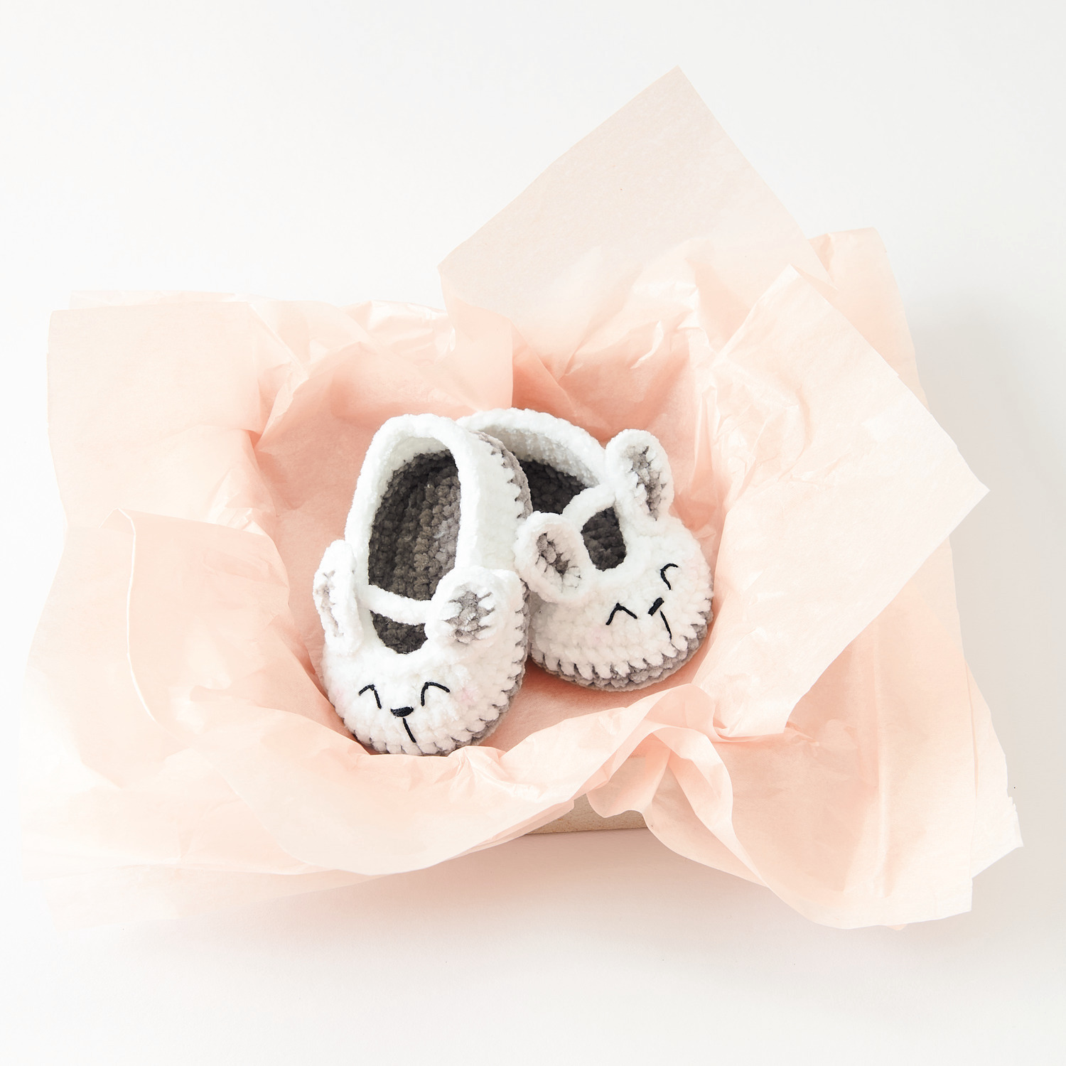Ricorumi Nilli Nilli Häkelset zeigt weiße Baby Booties in Hasenform auf rosa Papier. Ideal für Neugeborene.