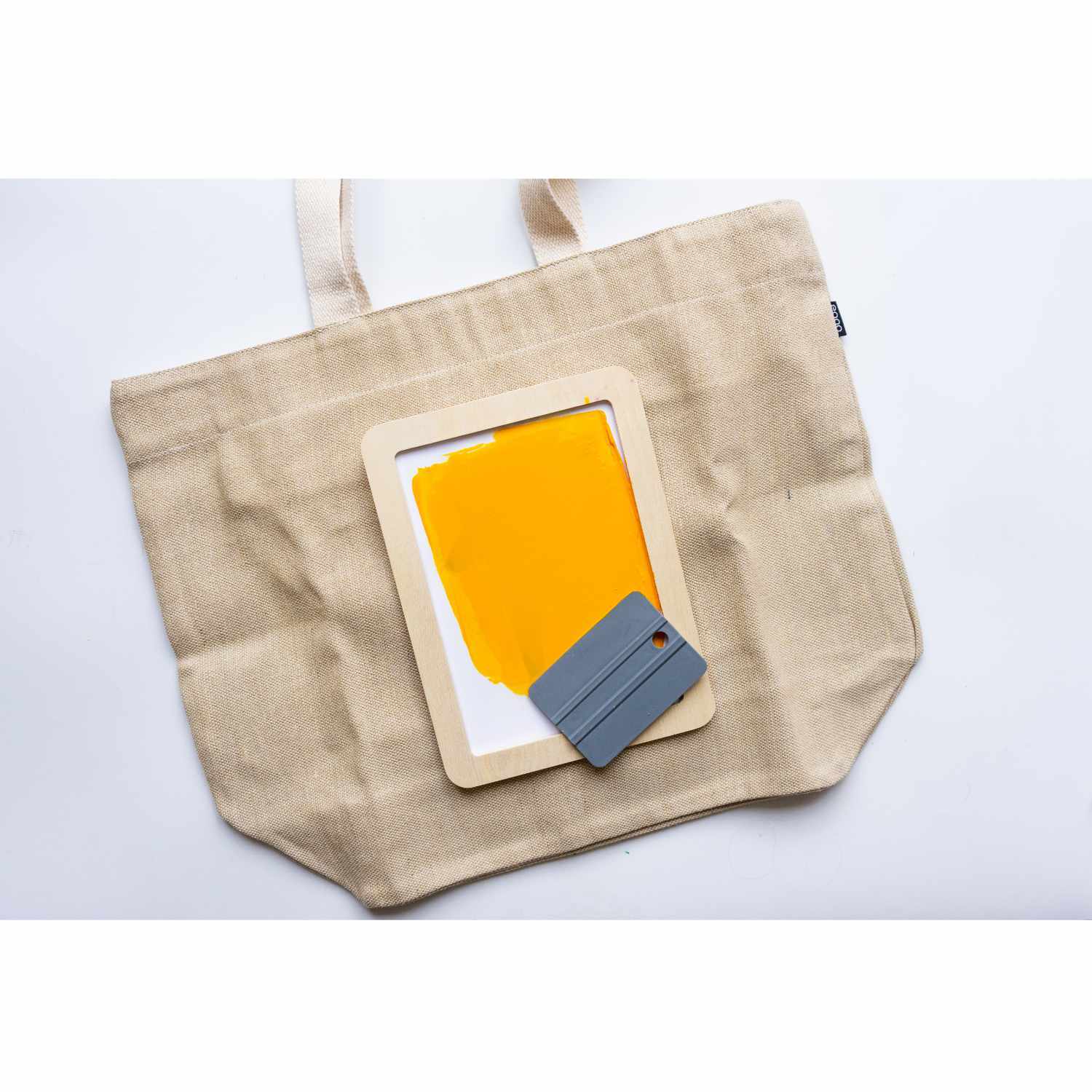 Textil Print Screen Siebdruck Set mit gelbem Druckrahmen und Rakel auf beiger Tasche. Ideal für kreative Textildruckprojekte.