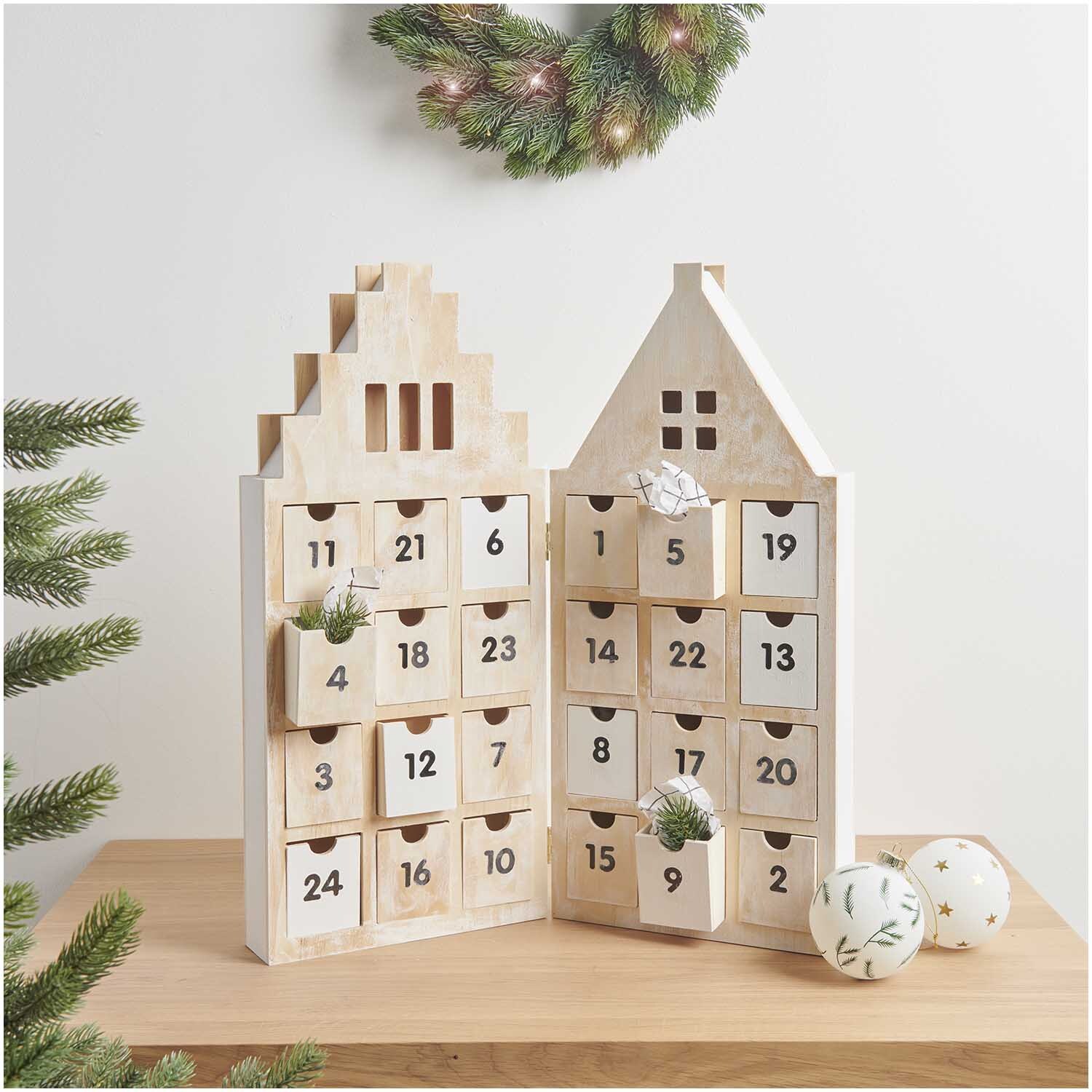 Adventskalender Haus aus Holz mit 24 nummerierten Schubladen, dekoriert mit Tannenzweigen und Kugeln, auf einem Tisch platziert.