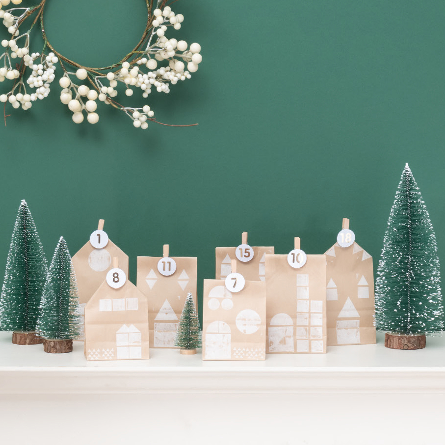 Bastelanleitung Adventskalender aus Papiertüten vor grünem Hintergrund mit Tannenbäumen und Beerenkranz.