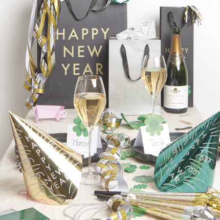 Gedeckter Tisch mit Tischkarten, Luftschlangen, Partyhüten, Geschenktüte und Sektgläsern. Fotosetting Tisch und weiße Wand.