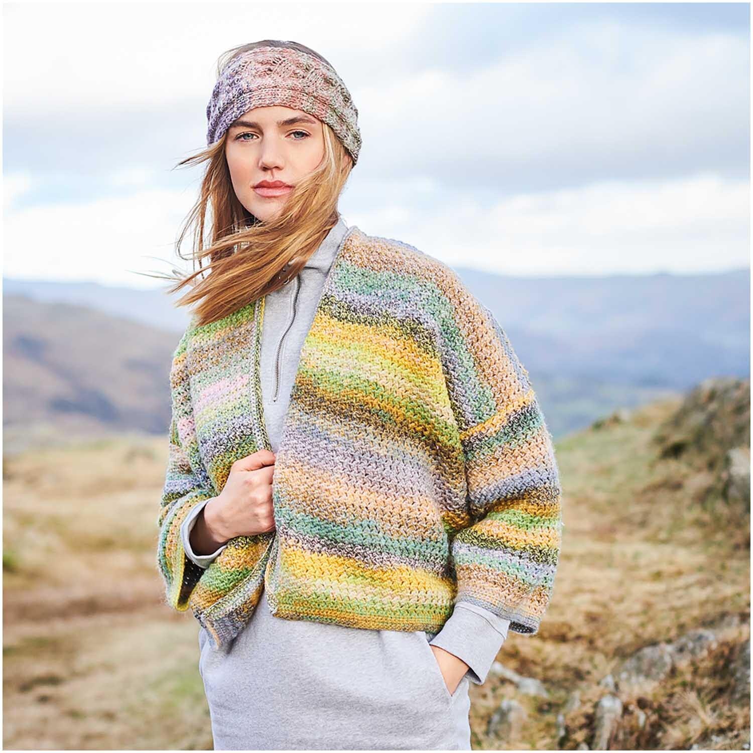 Häkelset Jacke Modell 05: Frau trägt bunte, gestrickte Jacke und Stirnband in natürlicher Landschaft.