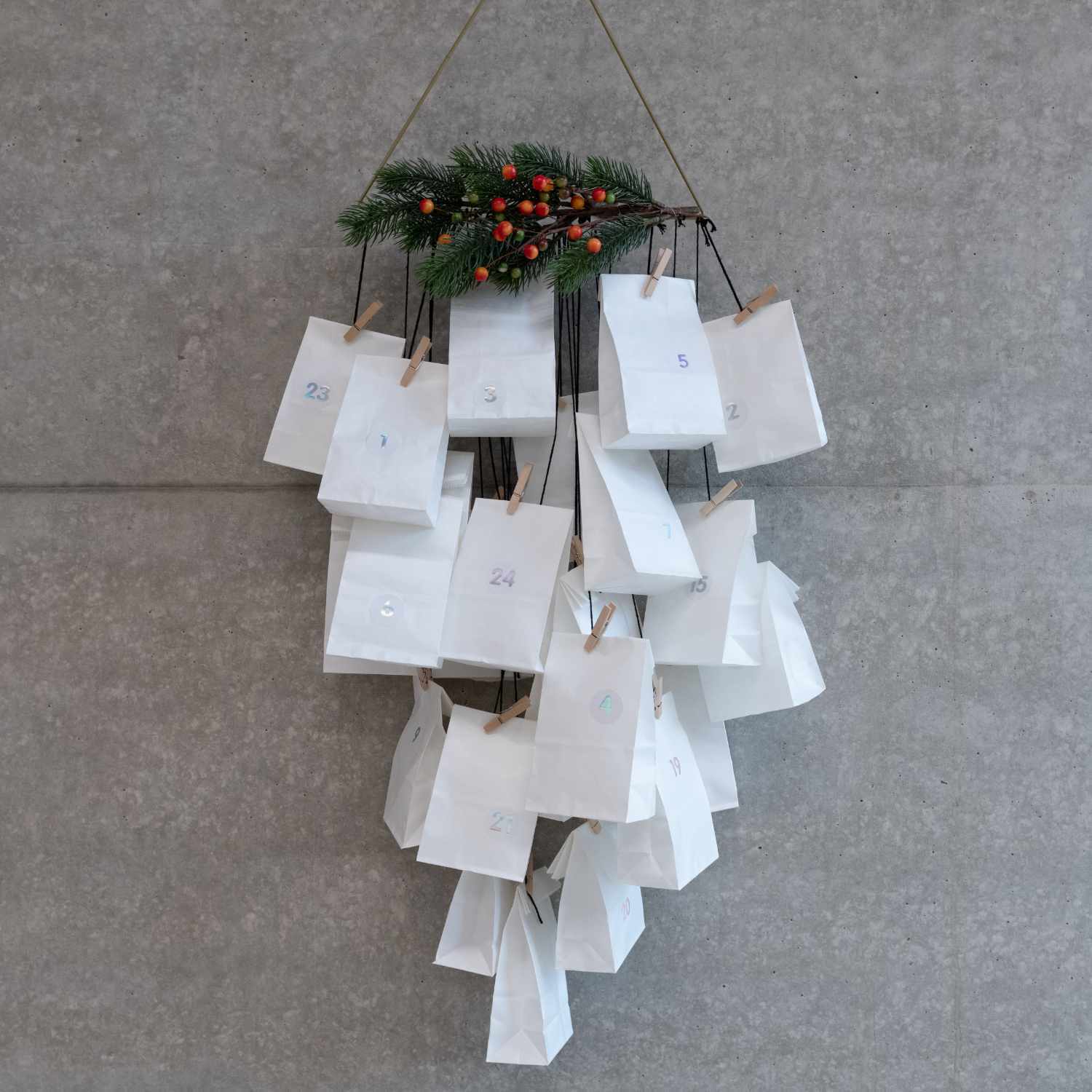 Bastelanleitung moderner Papiertüten-Adventskalender: Weiße Tüten mit Zahlen, dekoriert mit Tannenzweigen und Beeren, an einer Wand hängend.