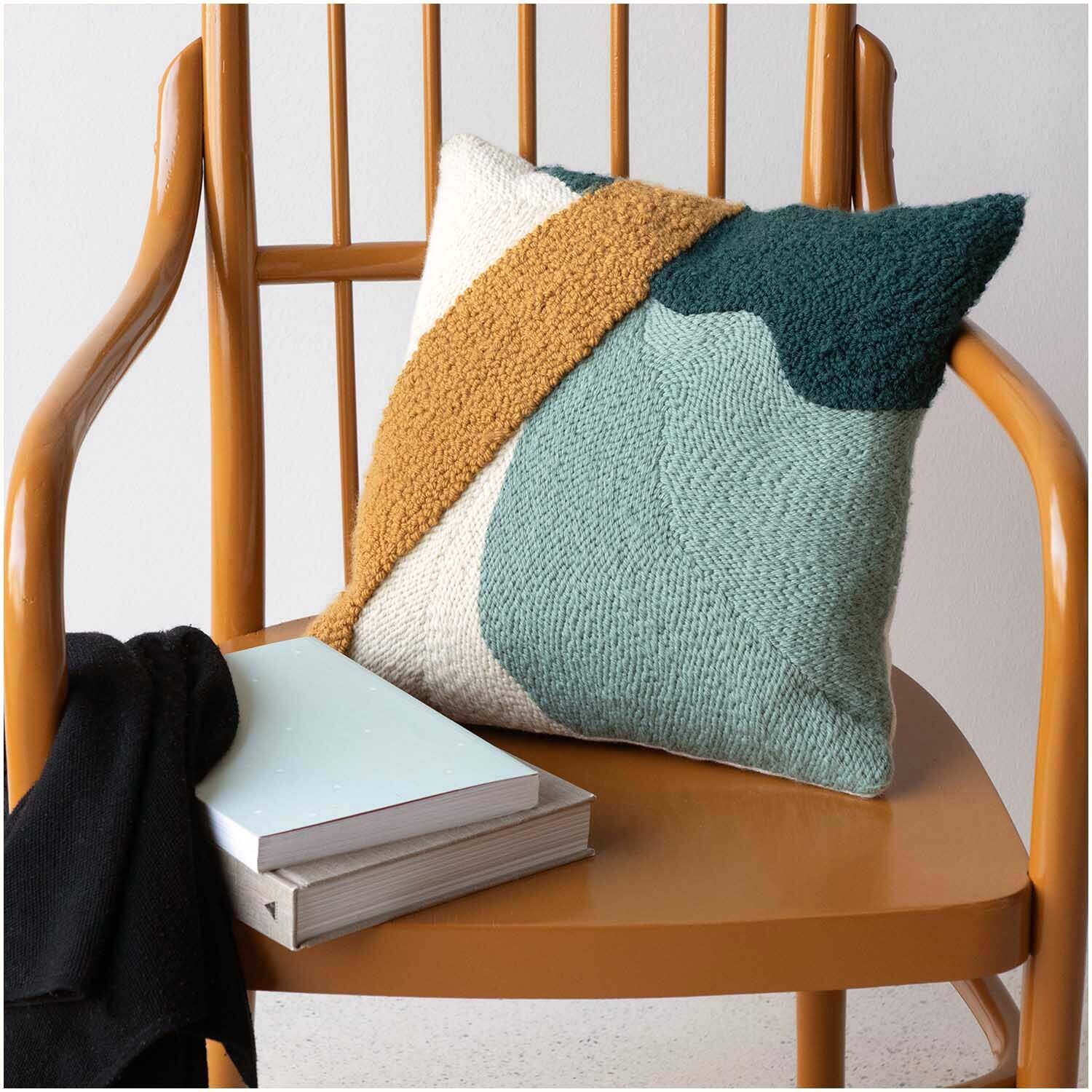 Punch Needle Packung Kissen in senf-grün auf Holzstuhl, dekoriert mit Büchern und Decke. Ideal für stilvolle Wohnräume.