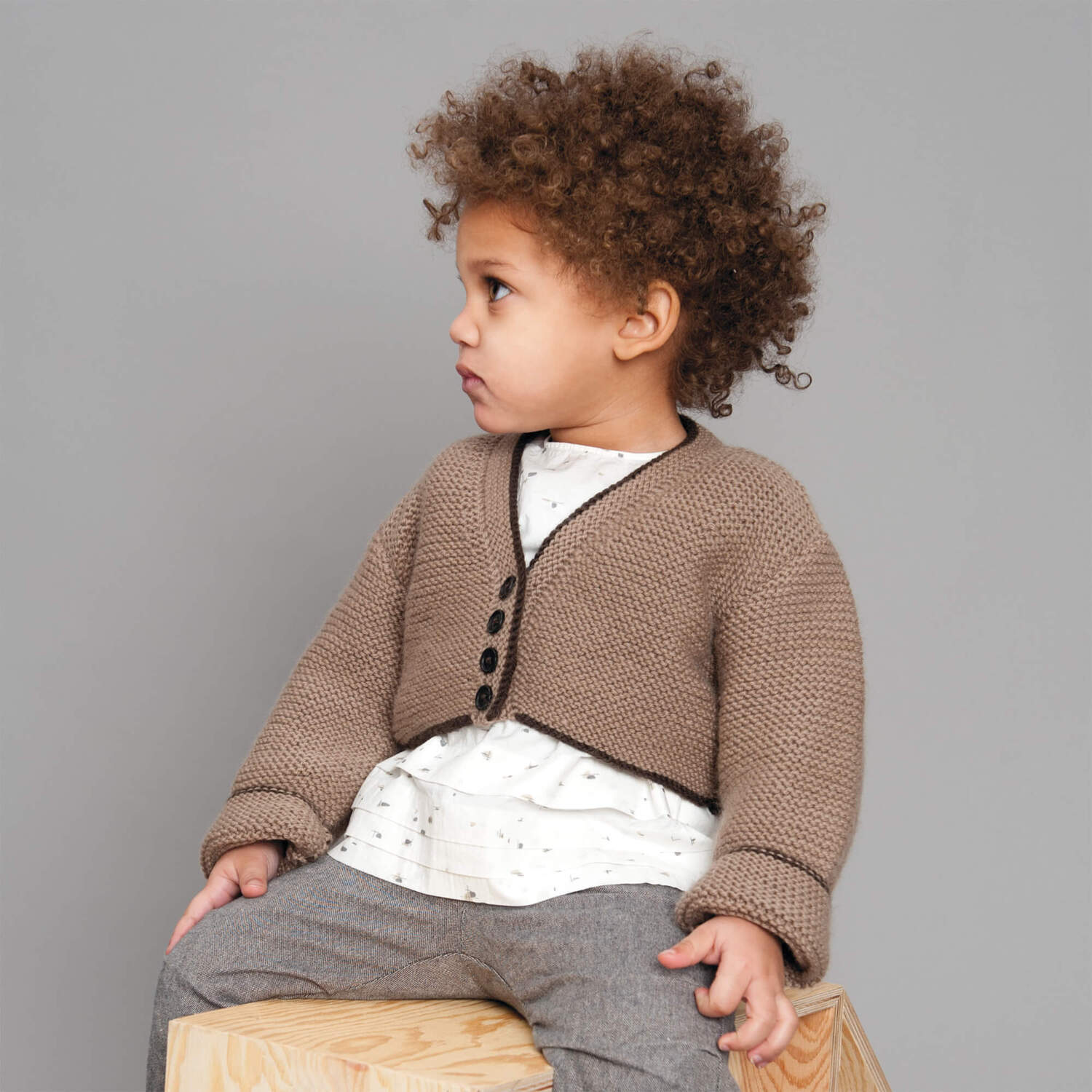 Strickanleitung Babybolero aus Essentials Cashlana: Kind trägt braunen Bolero mit Knöpfen, sitzt auf Holzblock, grauer Hintergrund.