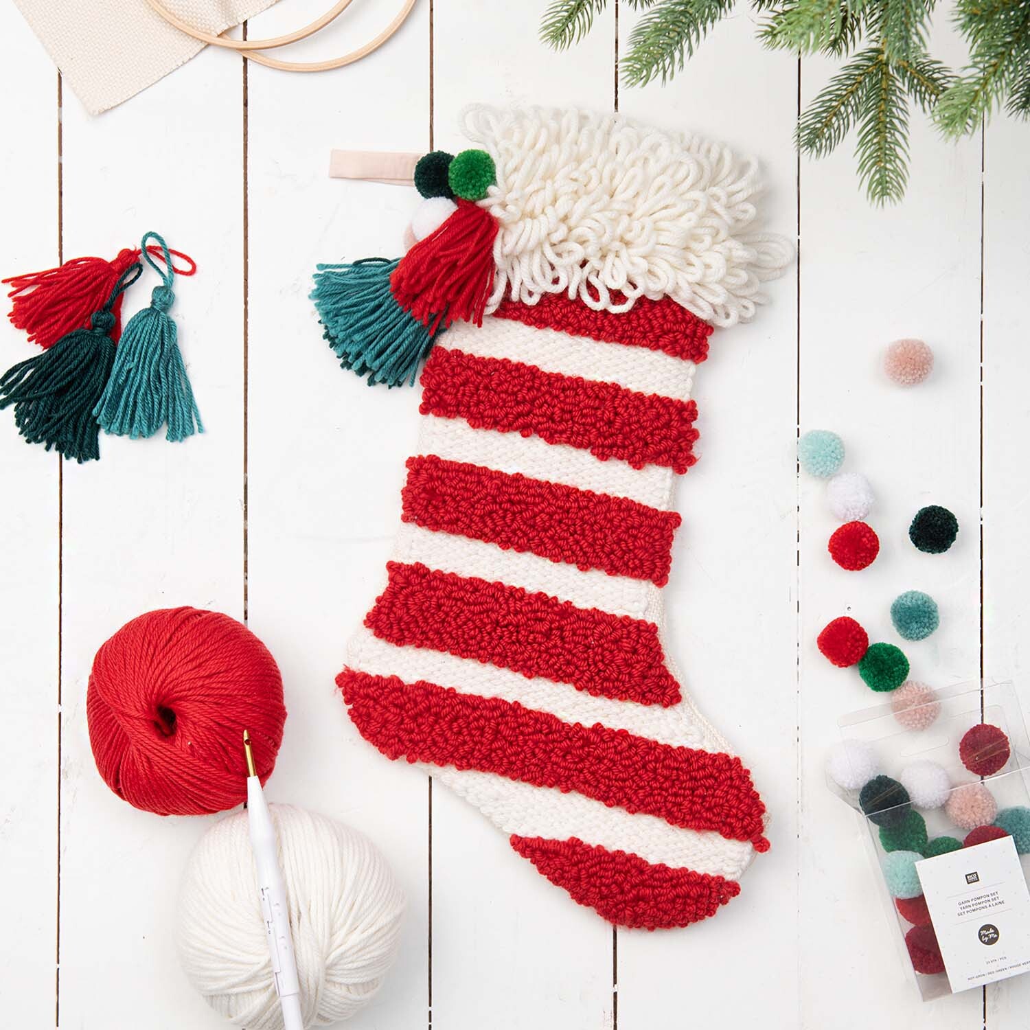 Anleitung gestreifter Punch Needle Weihnachtsstrumpf mit roten und weißen Streifen, umgeben von Garn und Pompons.