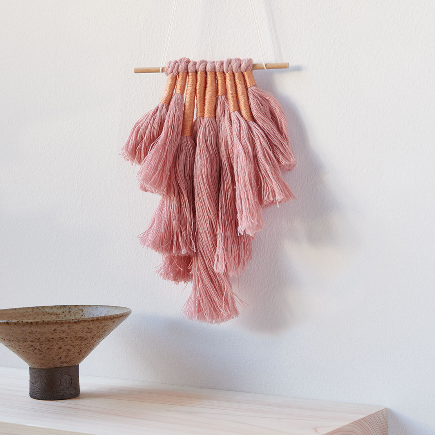 Bastelanleitung DIY Wandbehang in Erdtönen: Handgefertigter Wandbehang aus rosa Garn auf Holzstab, dekorativ an weißer Wand.