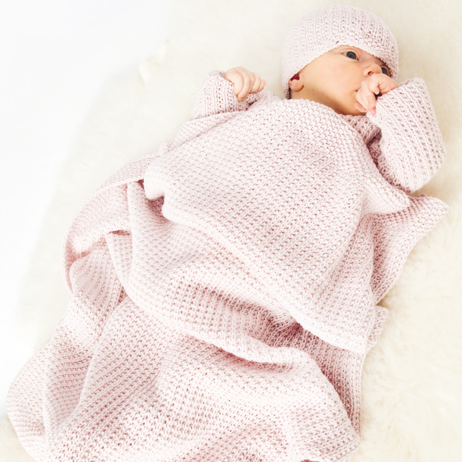 Strickanleitung Baby-Decke: Ein Baby liegt auf einer weichen, rosa gestrickten Decke und trägt eine passende Mütze.