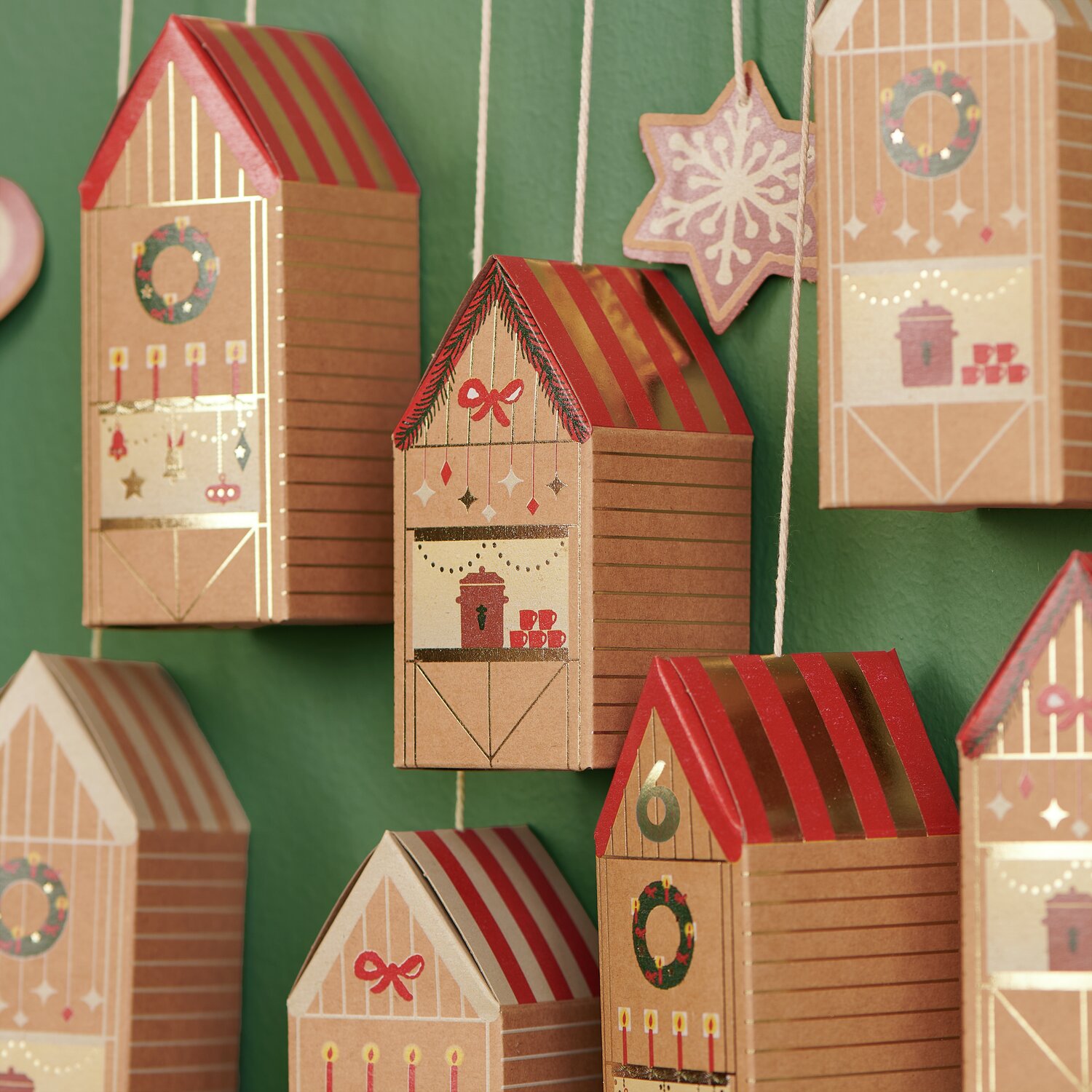 Adventskalender Bastelset: Hängende, festlich dekorierte Häuschen aus Karton in Rot und Braun vor grünem Hintergrund.