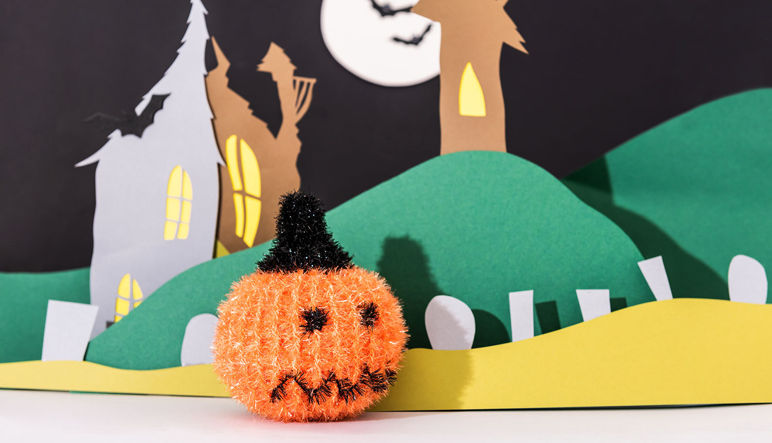 Häkelanleitung Creative Bubble Kürbis vor einer Halloween-Kulisse mit gruseligem Haus und Mond.