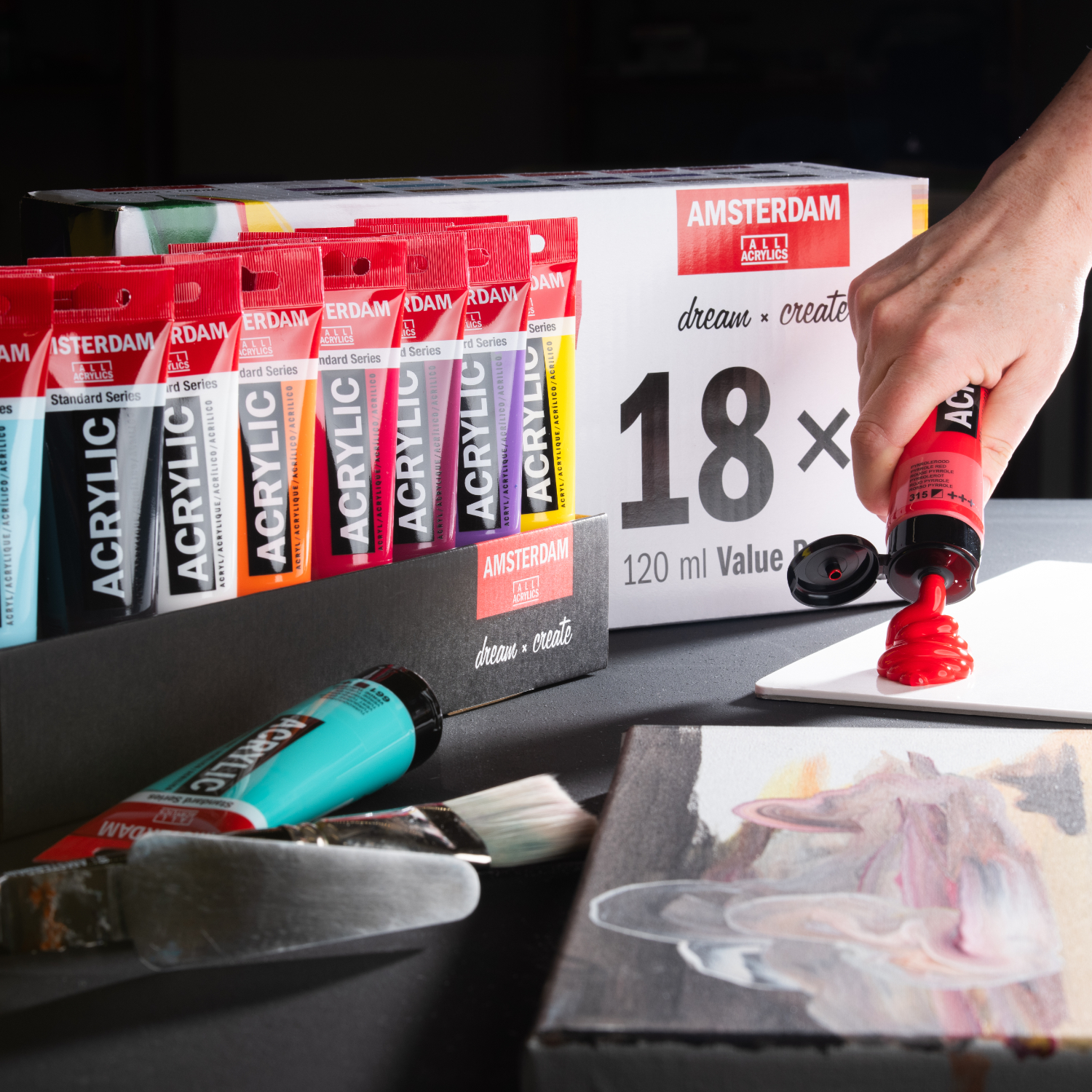 Vorteilspackung 18 x 120 ml: Eine Hand drückt rote Acrylfarbe aus einer Tube auf eine Palette. Im Hintergrund sind weitere bunte Farben der Marke AMSTERDAM zu sehen.