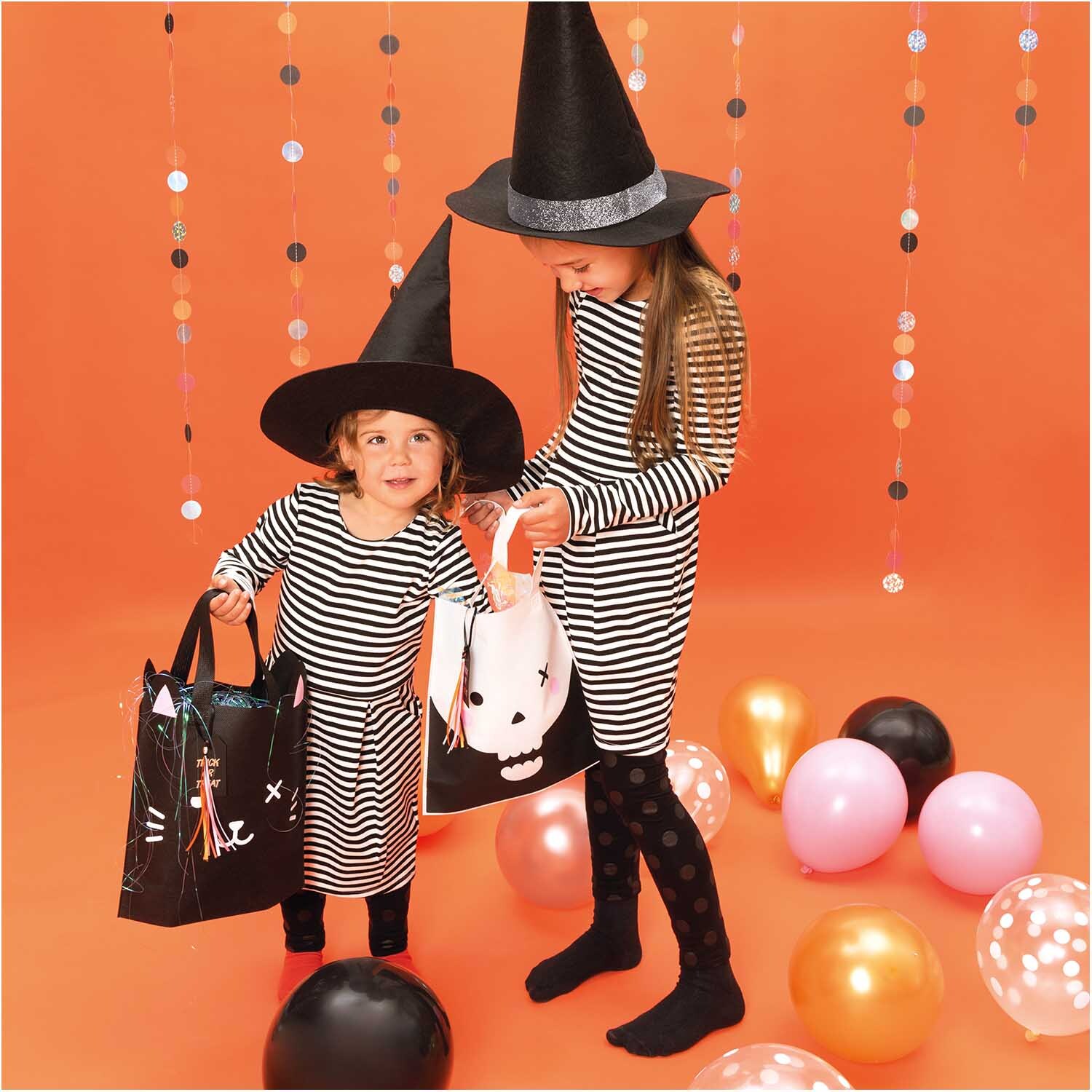 Luftballon Mix Halloween 30cm 12 Stück: Zwei Kinder in Hexenkostümen mit gestreiften Kleidern und Halloween-Taschen, umgeben von bunten Luftballons.