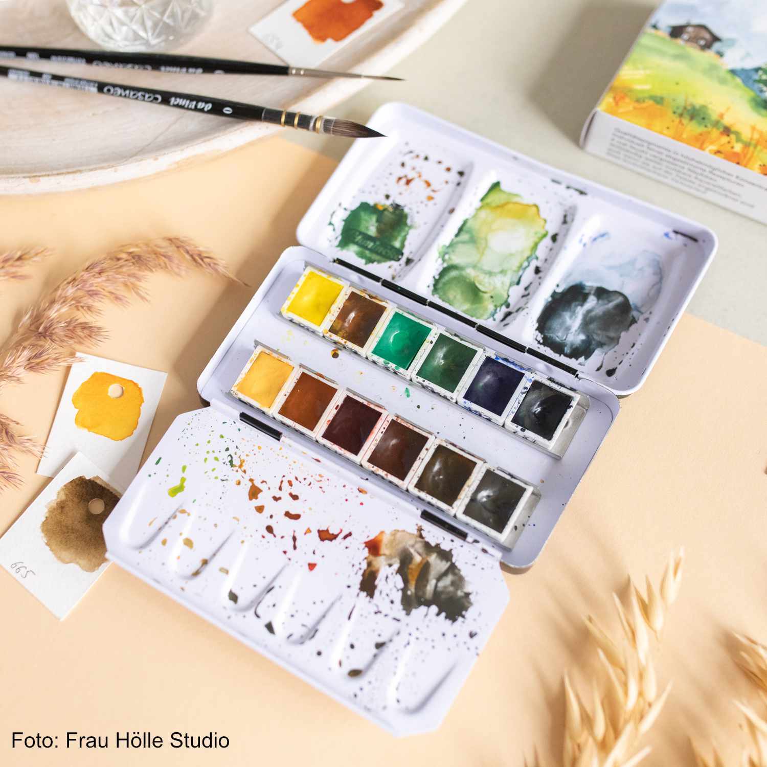 Schmincke Horadam Aquarellkasten mit 12 halben Näpfen, geöffnet, zeigt verschiedene Farbtöne. Pinsel und Farbproben daneben.