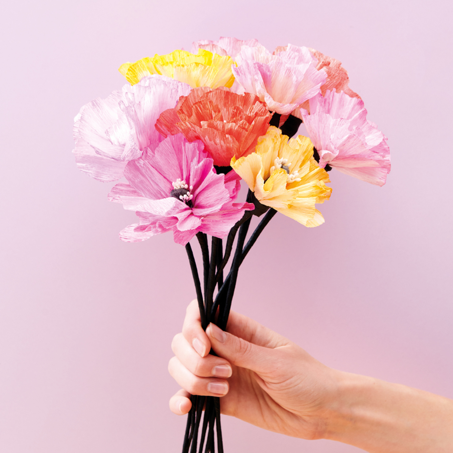 Bastelanleitung DIY-Papierblumen: Hand hält bunte Papierblumen vor einem rosa Hintergrund.