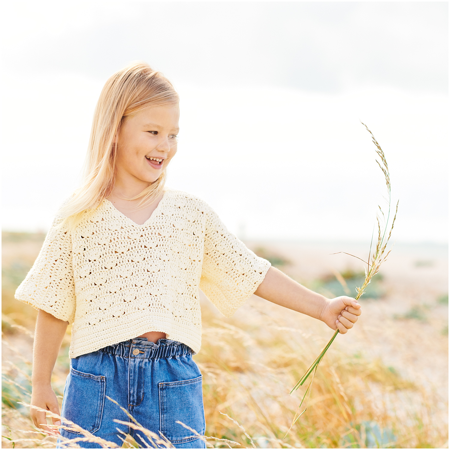 Essentials Organic Cotton dk: Mädchen in cremefarbenem Strickpullover von Rico Design auf Wiese.
