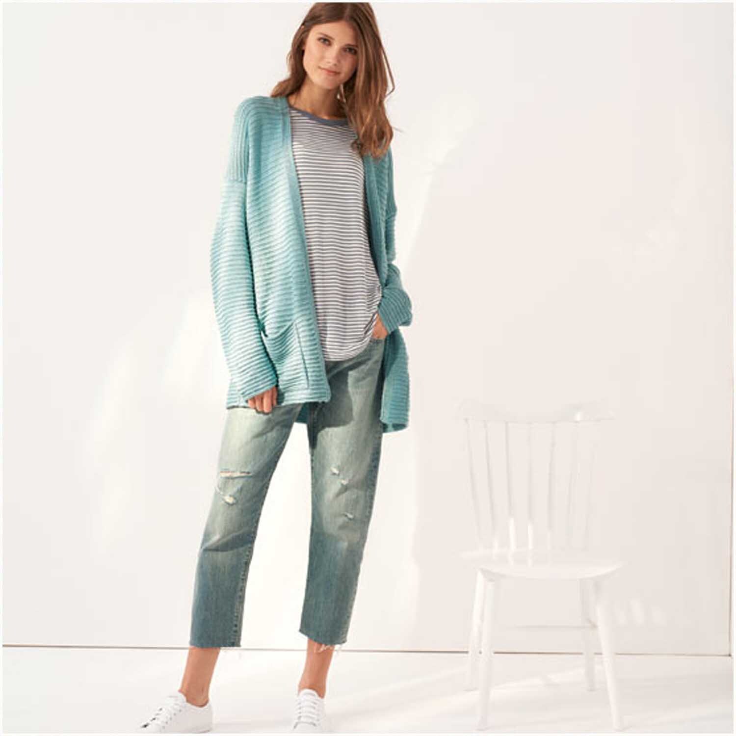 Creative Silky Touch dk: Frau in lässigem Outfit mit Aqua-Cardigan und Jeans, stehend neben einem weißen Stuhl. Ideal für modische Freizeitlooks.